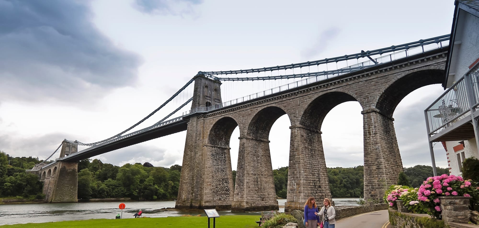 A large stone suspension bridge with multiple arches spans a river, with two people walking on a path below and greenery in the background.